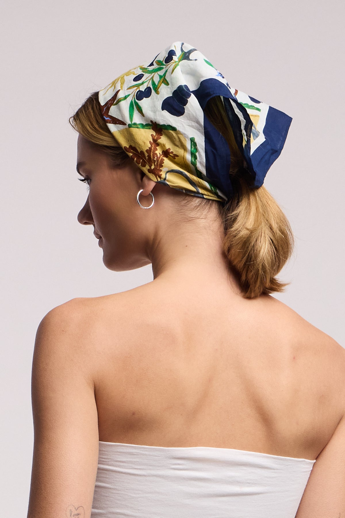 A woman with light brown hair wears the ZURI SCARF by Velvet by Graham & Spencer as a headscarf and silver hoop earrings, facing away in a white strapless top against a plain background.