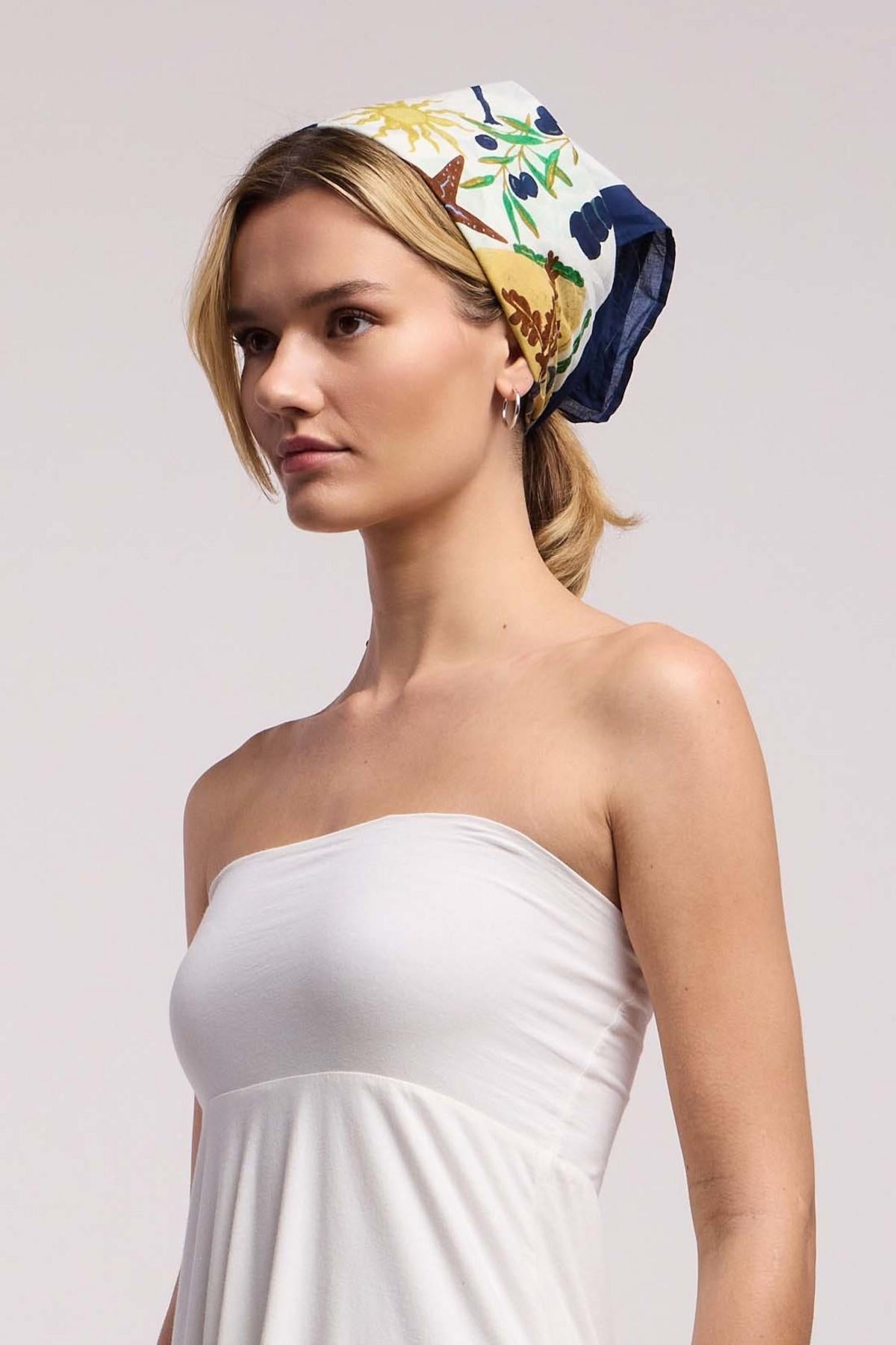 A woman stands against a plain background, wearing a white strapless dress and the ZURI SCARF by Velvet by Graham & Spencer as a colorful headscarf.-41548249530561