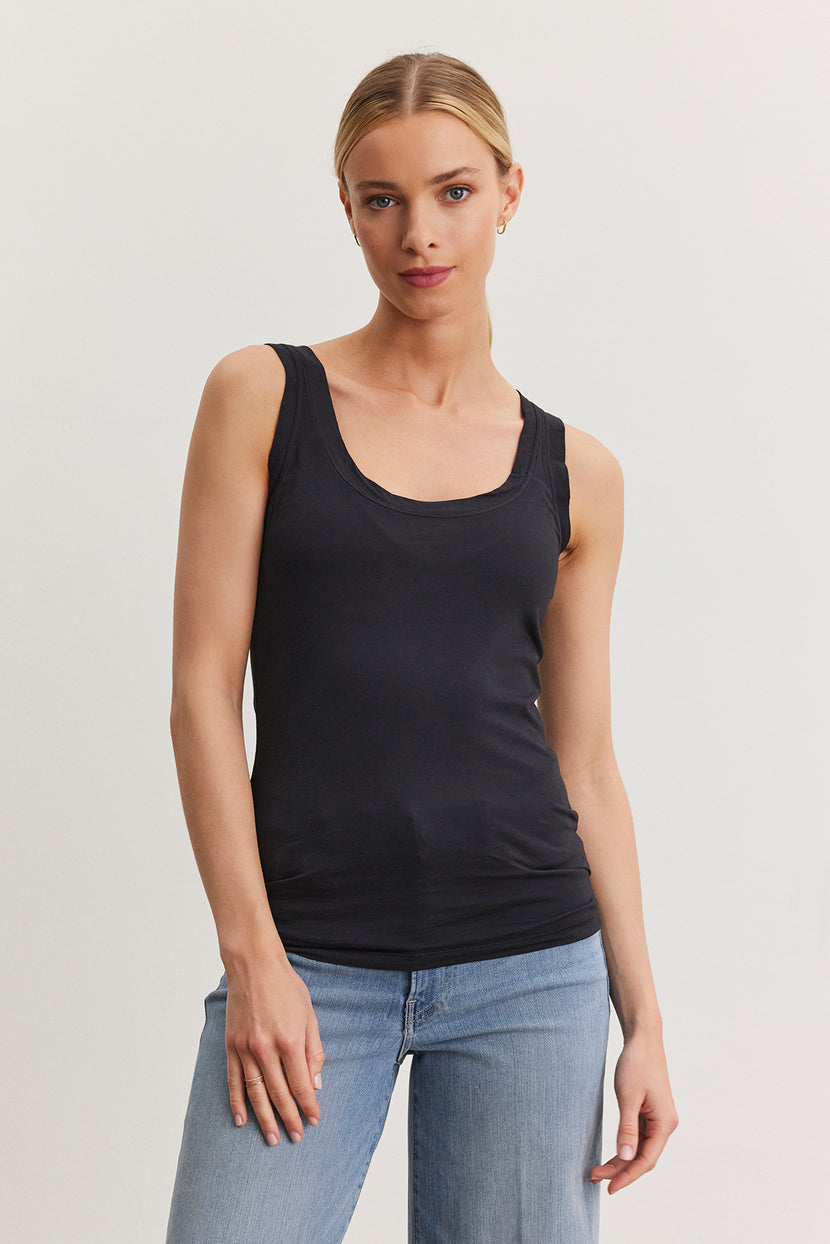 A person stands against a plain white background wearing a fitted MOSSY TANK TOP by Velvet by Graham & Spencer and blue jeans, showcasing the versatility of their wardrobe.