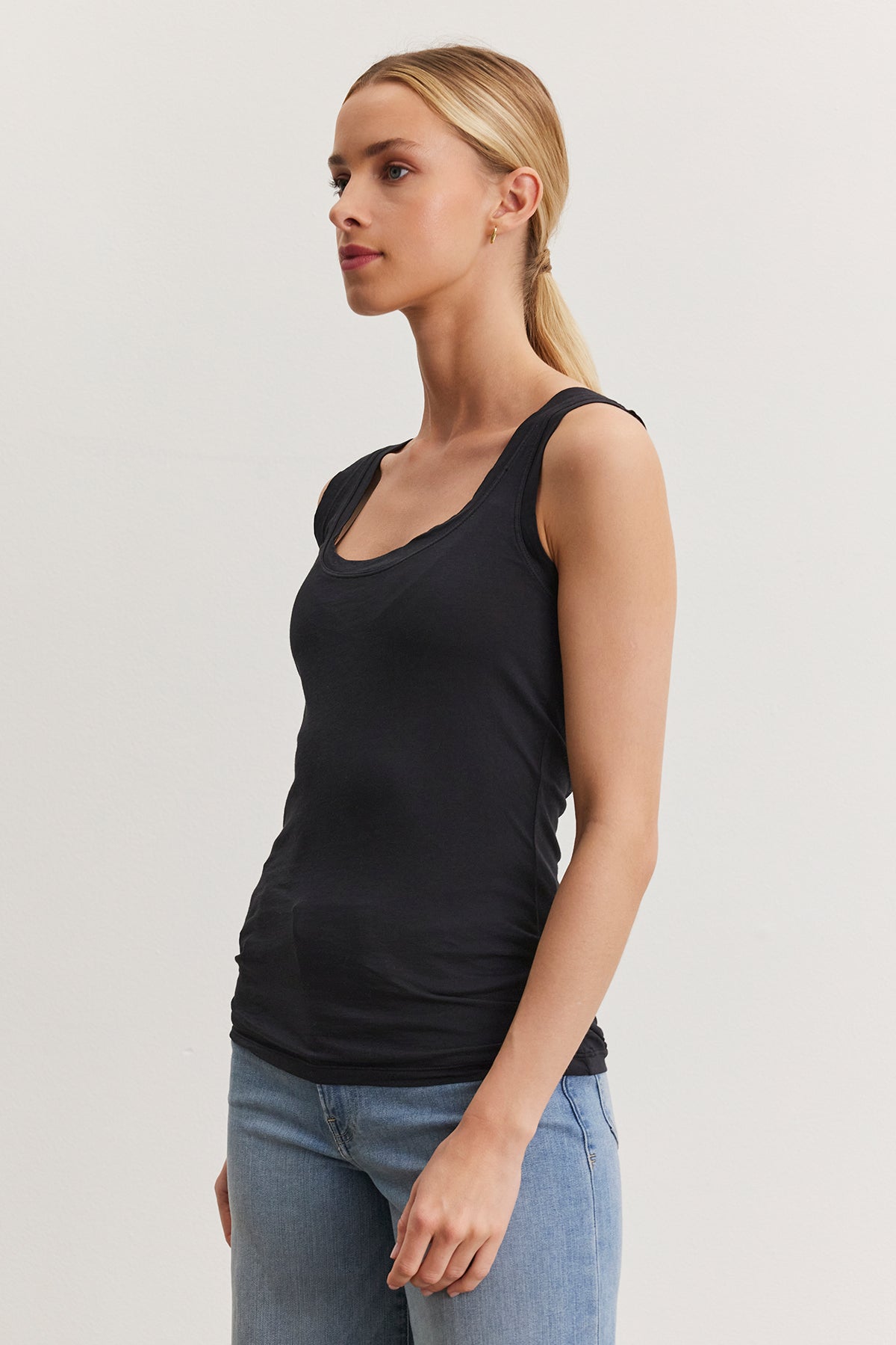 A person with light brown hair in a ponytail is wearing a black MOSSY TANK TOP by Velvet by Graham & Spencer and blue jeans, standing against a plain white background and looking to the side.-36984053006529