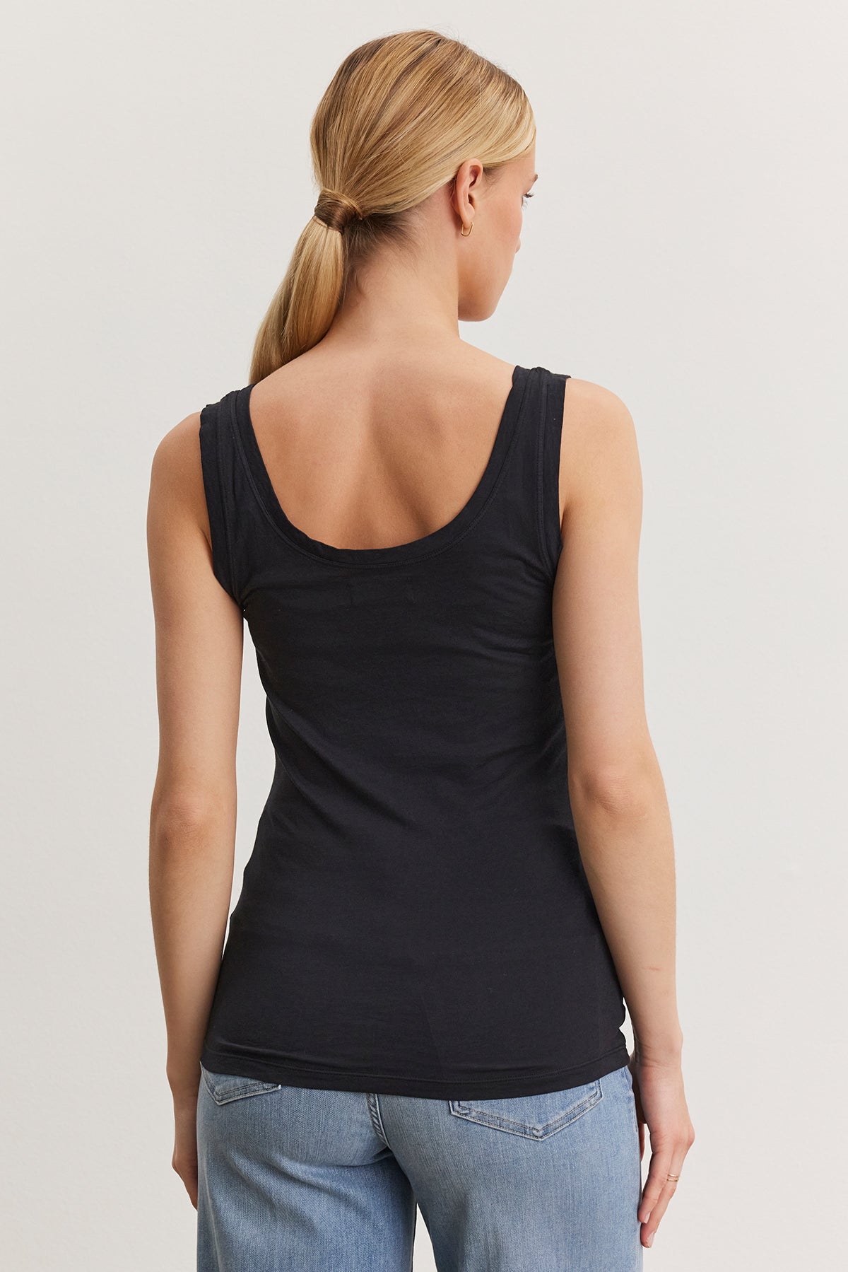 A woman with blonde hair in a low ponytail, wearing a fitted black MOSSY TANK TOP by Velvet by Graham & Spencer and light blue jeans, is viewed from the back against a plain white background.-36984053039297
