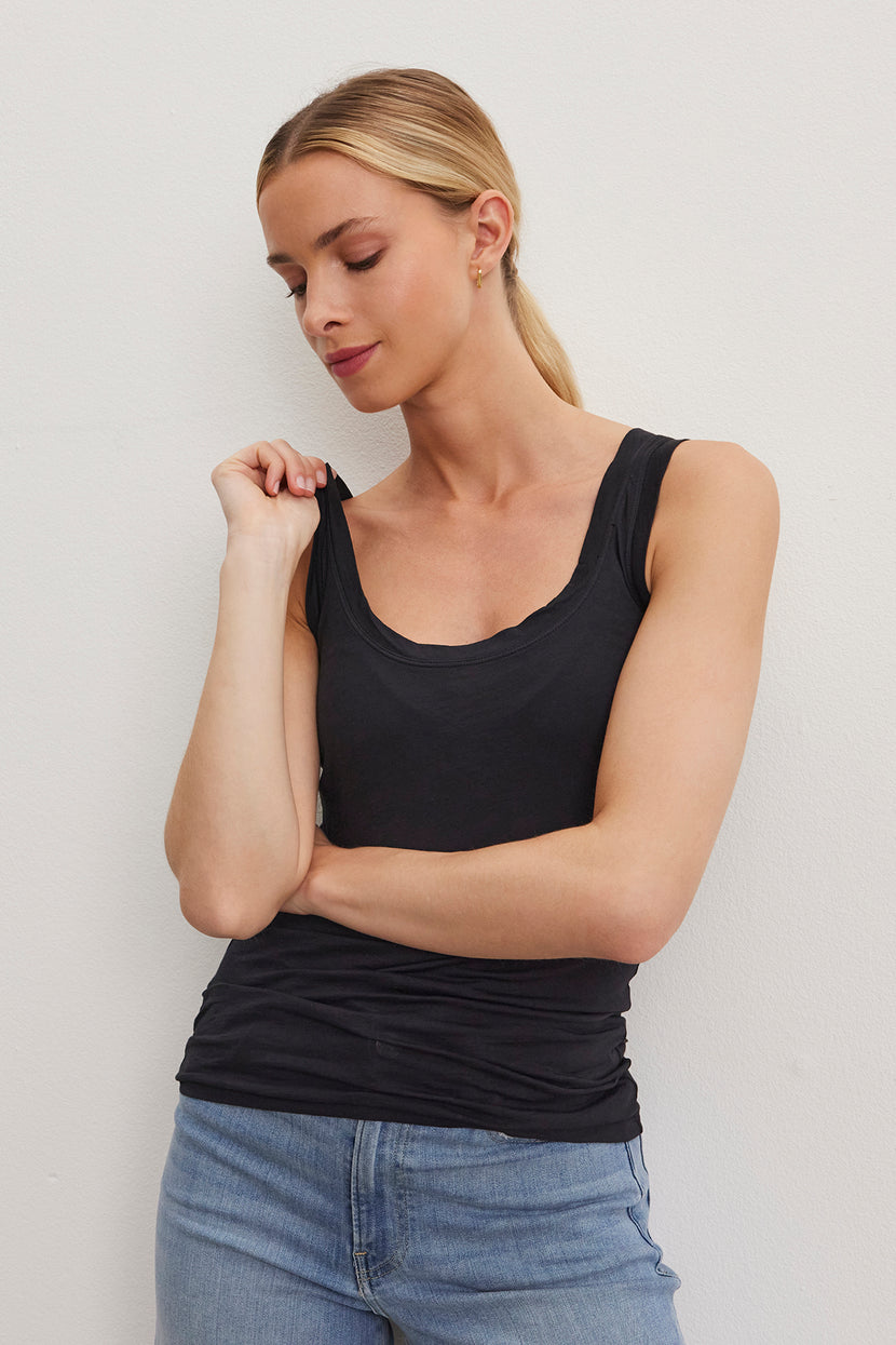 A woman with blonde hair, wearing the stylish MOSSY TANK TOP from Velvet by Graham & Spencer and blue jeans, stands against a plain white background. She looks downward with her left hand touching her shoulder and right arm crossed over her torso, effortlessly showcasing the versatility of a well-layered wardrobe.