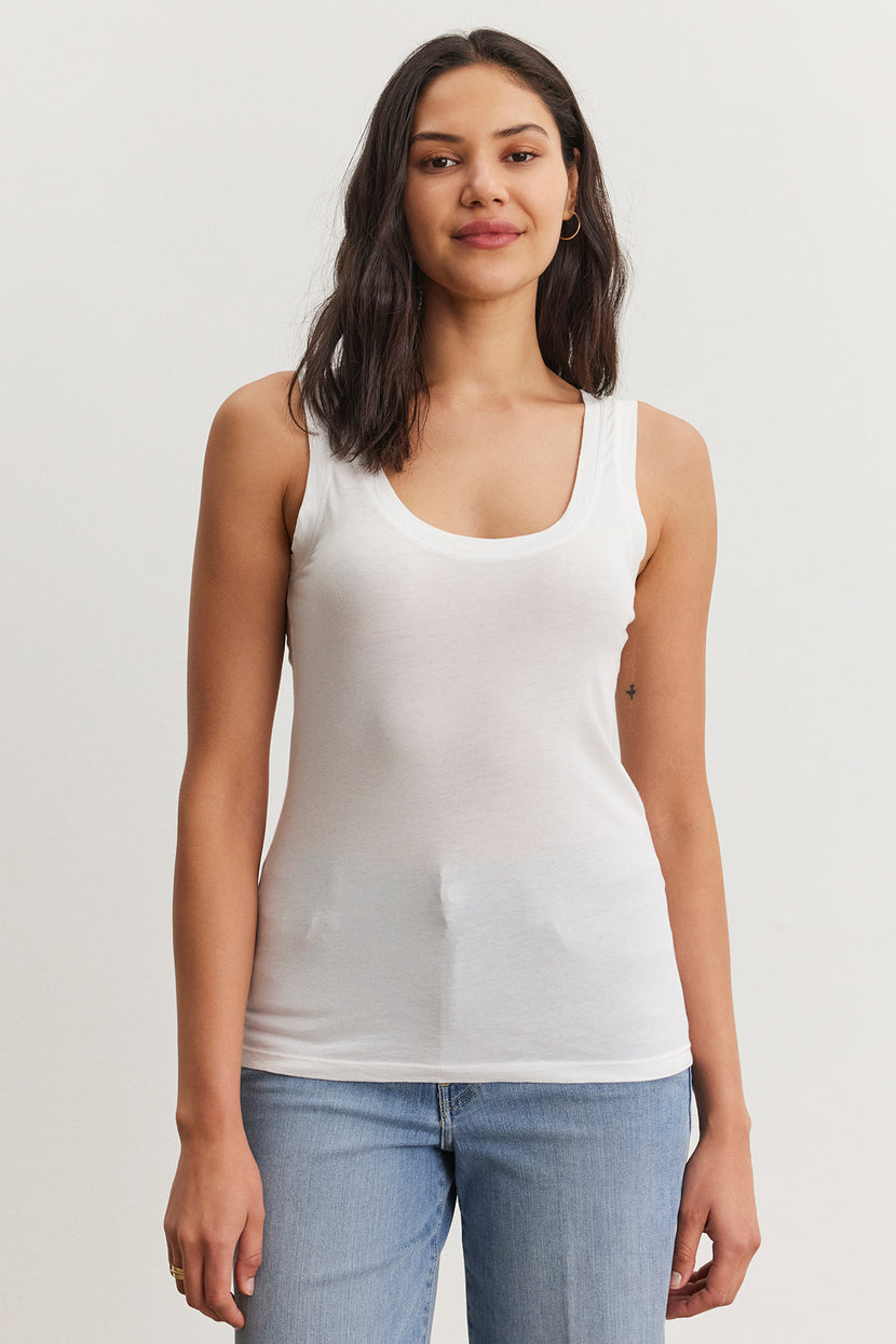 A person with shoulder-length dark hair is standing, wearing a fitted MOSSY TANK TOP by Velvet by Graham & Spencer and blue jeans, showcasing a versatile wardrobe against a plain white background.