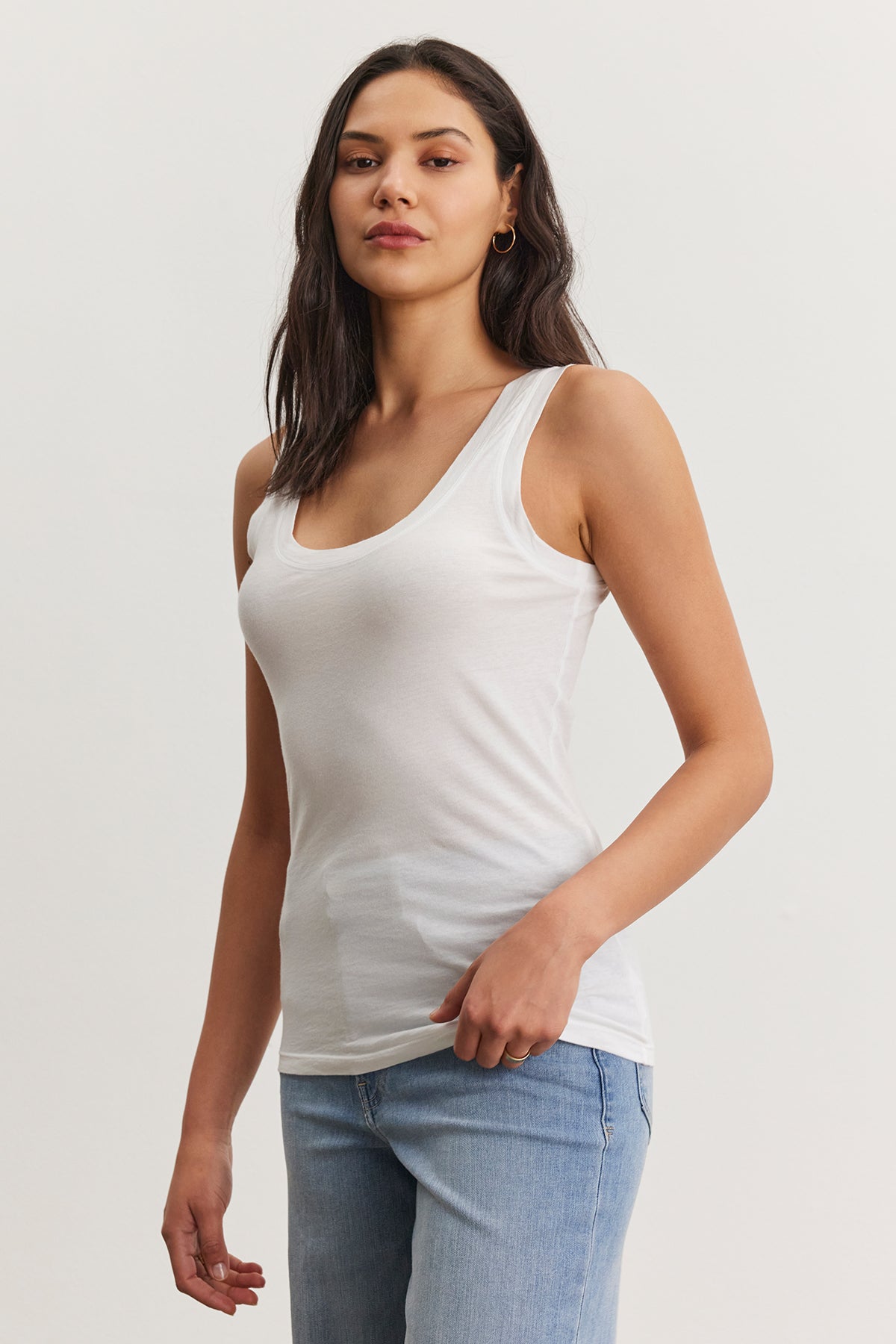   A person with long dark hair is wearing a white, fitted MOSSY TANK TOP by Velvet by Graham & Spencer and light blue jeans, standing with a neutral expression against a plain background. This ensemble highlights the versatility of a well-curated wardrobe. 