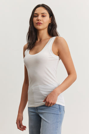 A person with long dark hair is wearing a white, fitted MOSSY TANK TOP by Velvet by Graham & Spencer and light blue jeans, standing with a neutral expression against a plain background. This ensemble highlights the versatility of a well-curated wardrobe.