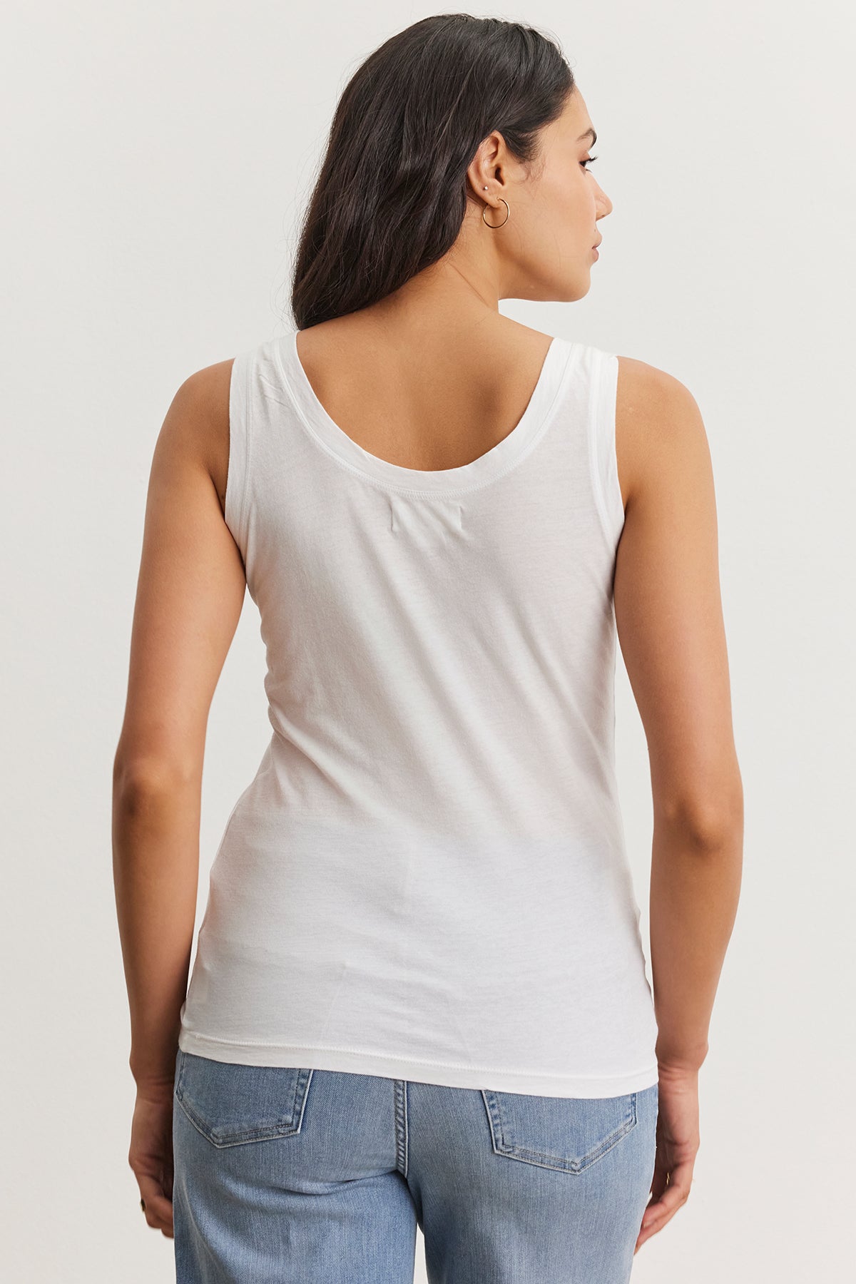   A woman with long dark hair is standing with her back to the camera, wearing a white fitted MOSSY TANK TOP by Velvet by Graham & Spencer and blue jeans, showcasing a versatile wardrobe. 