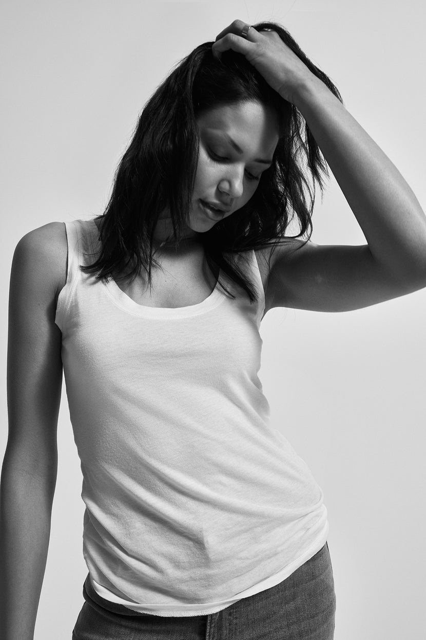 Black and white photo of a person with dark hair, wearing the MOSSY TANK TOP from Velvet by Graham & Spencer and jeans, looking down while resting a hand on their head. The fitted silhouette creates a timeless look that serves as a versatile wardrobe addition.