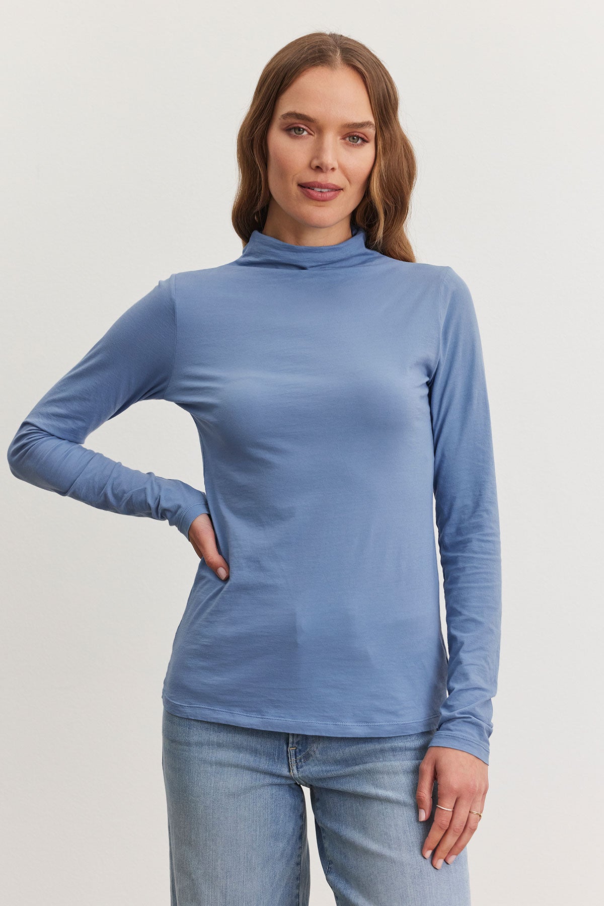   Against a plain background, a woman models the elegant TALISIA TEE by Velvet by Graham & Spencer, paired with jeans to create an ideal versatile wardrobe staple. With her right hand on her hip and long, loose hair flowing freely, she exudes effortless flair. 