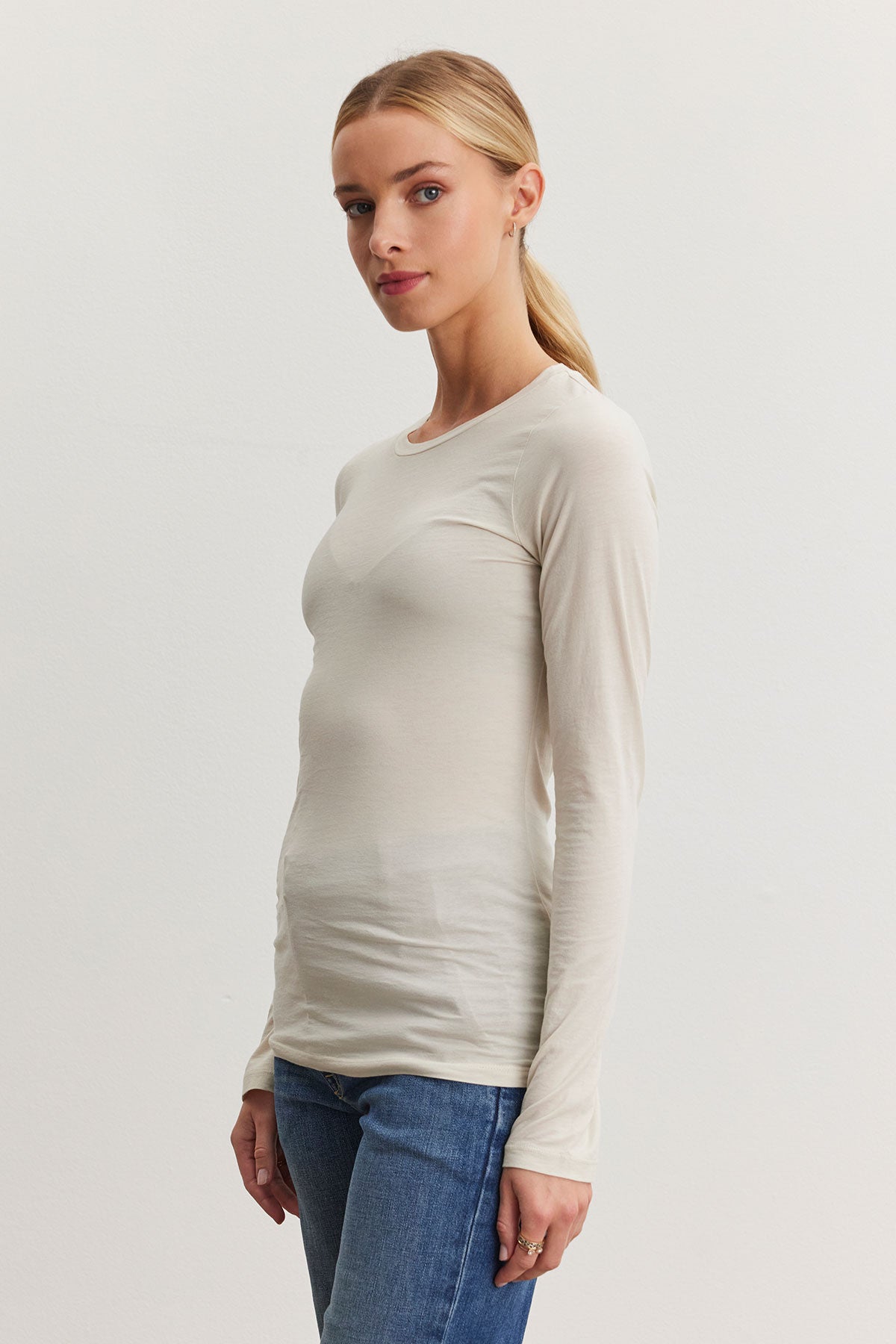   A person stands against a plain background wearing the ultra-soft ZOFINA TEE by Velvet by Graham & Spencer, a long-sleeve white shirt with a classic crew neckline, paired with blue jeans, their hair tied back. 