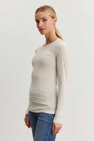 A person stands against a plain background wearing the ultra-soft ZOFINA TEE by Velvet by Graham & Spencer, a long-sleeve white shirt with a classic crew neckline, paired with blue jeans, their hair tied back.