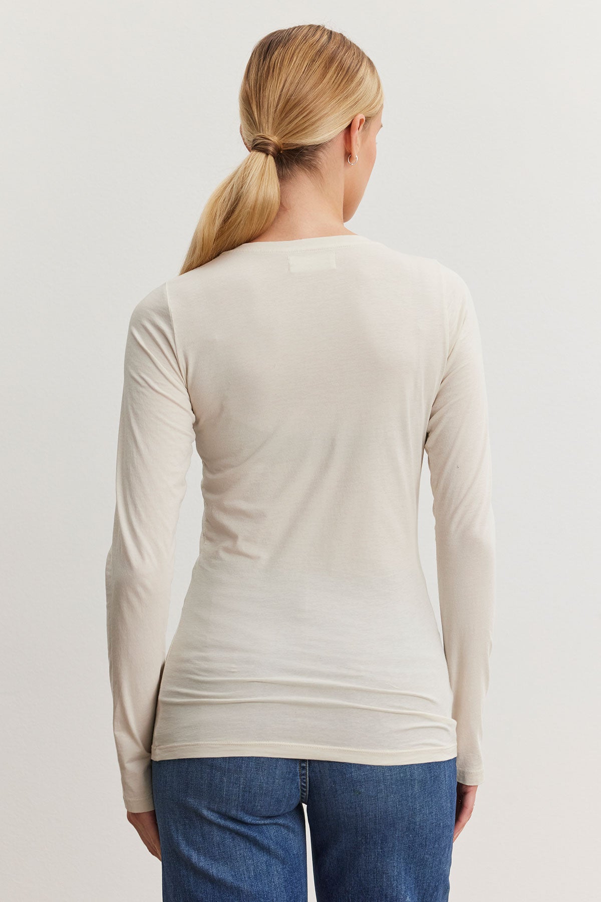   A woman with long blond hair in a ponytail is wearing the ZOFINA TEE by Velvet by Graham & Spencer, an ultra-soft gauzy whisper cream shirt with a classic crew neckline, paired with jeans, as she stands with her back to the camera. 