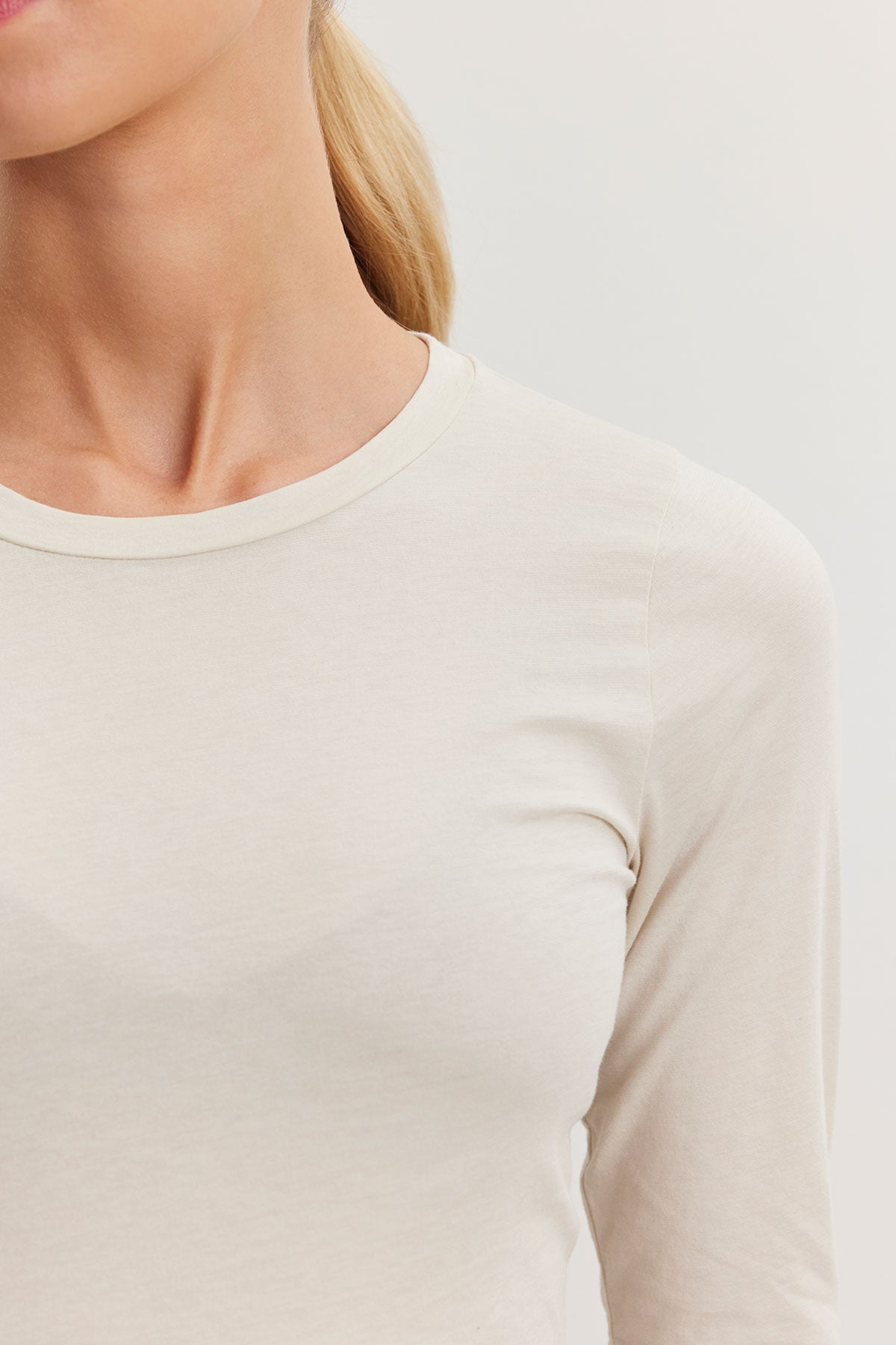   A person wearing the ZOFINA TEE by Velvet by Graham & Spencer—an ultra-soft gauzy whisper beige fitted long sleeve tee with a classic crew neckline—has her blonde hair tied back neatly against a neutral background. 