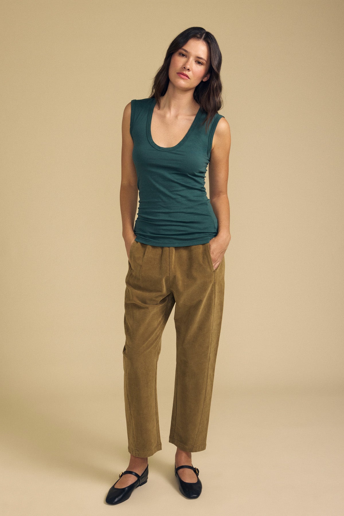 A woman stands against a beige background, wearing the Velvet by Graham & Spencer ESTINA TANK TOP—a sleeveless dark green scoop-neck tank—paired with tan high-waisted pants and black shoes, her hands in her pockets.