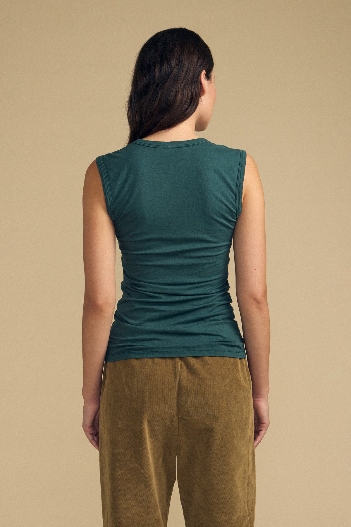 A person stands facing away from the camera, wearing the Velvet by Graham & Spencer ESTINA TANK TOP in a soft gauzy green fabric paired with brown pants, set against a plain beige background.