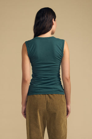 A person stands facing away from the camera, wearing the Velvet by Graham & Spencer ESTINA TANK TOP in a soft gauzy green fabric paired with brown pants, set against a plain beige background.