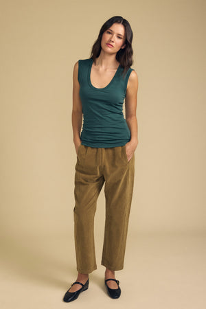 A woman stands against a beige background, wearing the Velvet by Graham & Spencer ESTINA TANK TOP—a sleeveless dark green scoop-neck tank—paired with tan high-waisted pants and black shoes, her hands in her pockets.