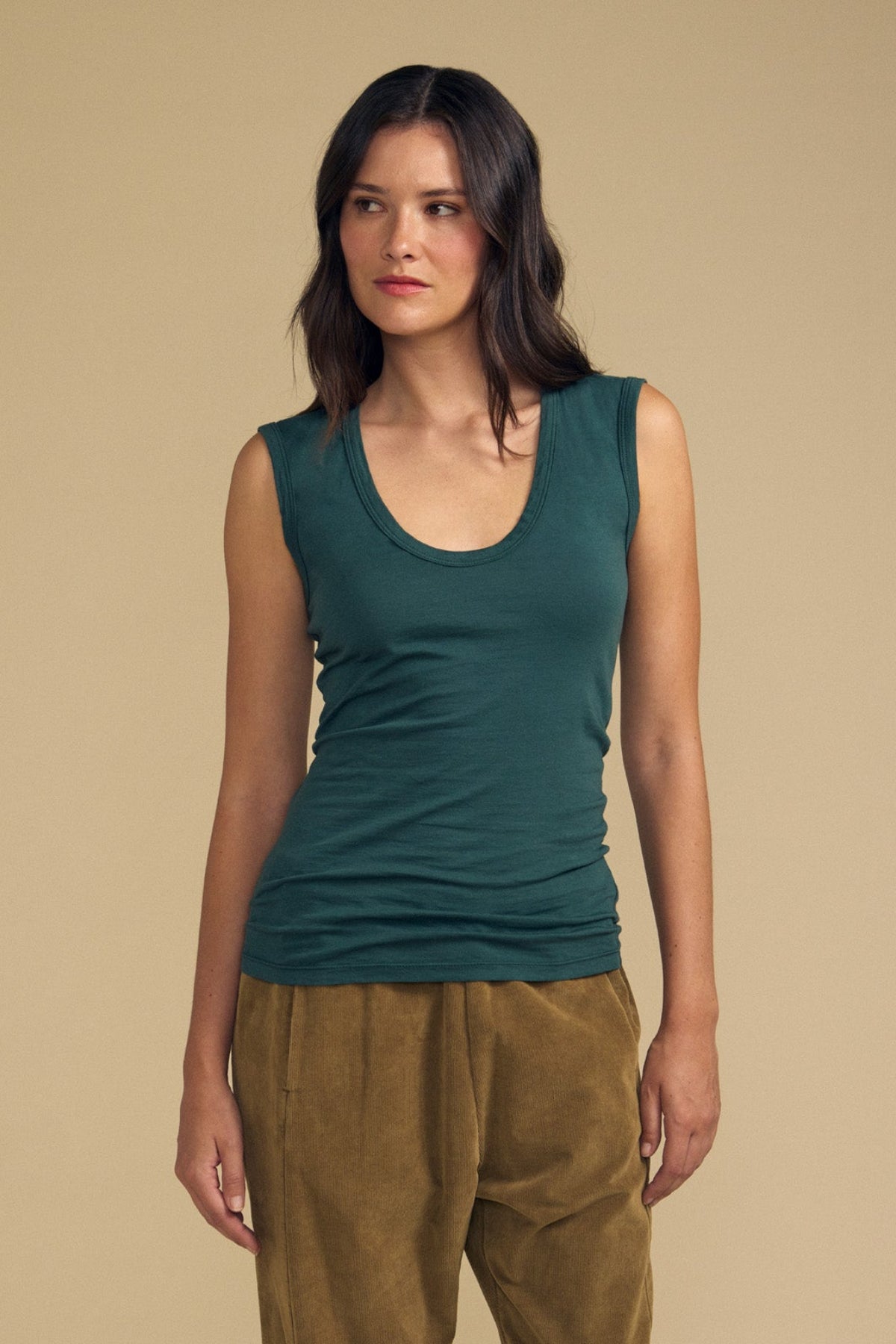 A woman stands against a beige background, wearing the ESTINA TANK TOP in soft gauzy whisper green by Velvet by Graham & Spencer and brown pants, looking to her left with a neutral expression.-41815071490241