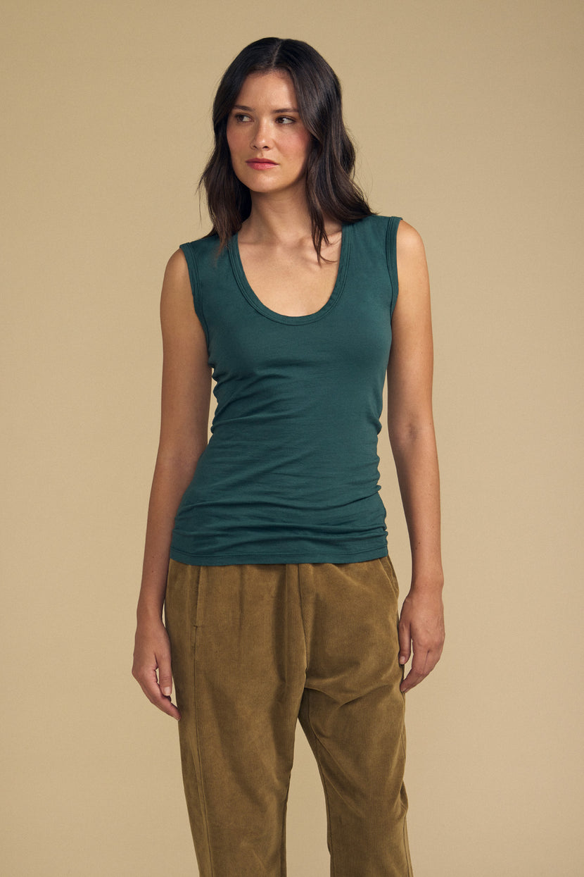 A woman stands against a beige background, wearing the ESTINA TANK TOP in soft gauzy whisper green by Velvet by Graham & Spencer and brown pants, looking to her left with a neutral expression.