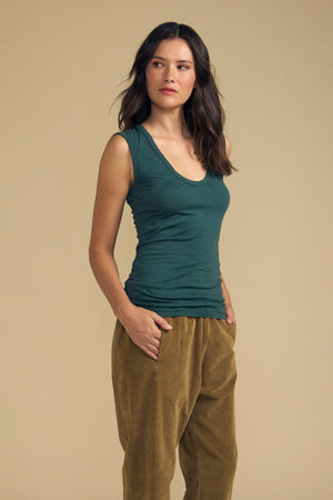 A woman stands against a beige background, wearing the ESTINA TANK TOP in green by Velvet by Graham & Spencer paired with brown pants, one hand in her pocket as she looks slightly to the side.