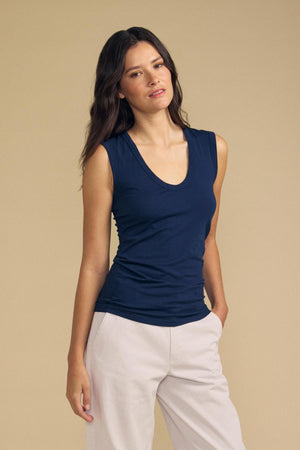 Wearing the ESTINA TANK TOP by Velvet by Graham & Spencer in a relaxed sleeveless navy style, a person with long brown hair pairs it with light beige pants, standing against a plain beige background.