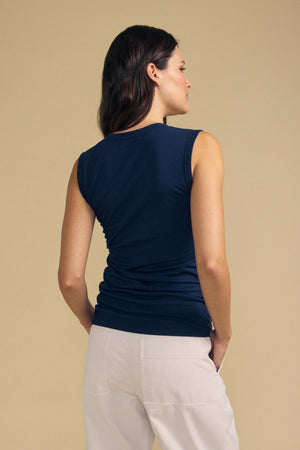 A woman with long brown hair stands facing away against a beige background, wearing the ESTINA TANK TOP by Velvet by Graham & Spencer—a navy scoop-neck laid-back tank—paired with light-colored pants.