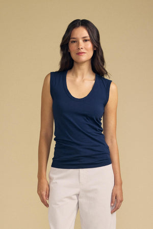 A woman wearing the ESTINA TANK TOP by Velvet by Graham & Spencer—a navy, laid-back scoop-neck tank—paired with light-colored pants stands against a plain beige background, facing the camera.