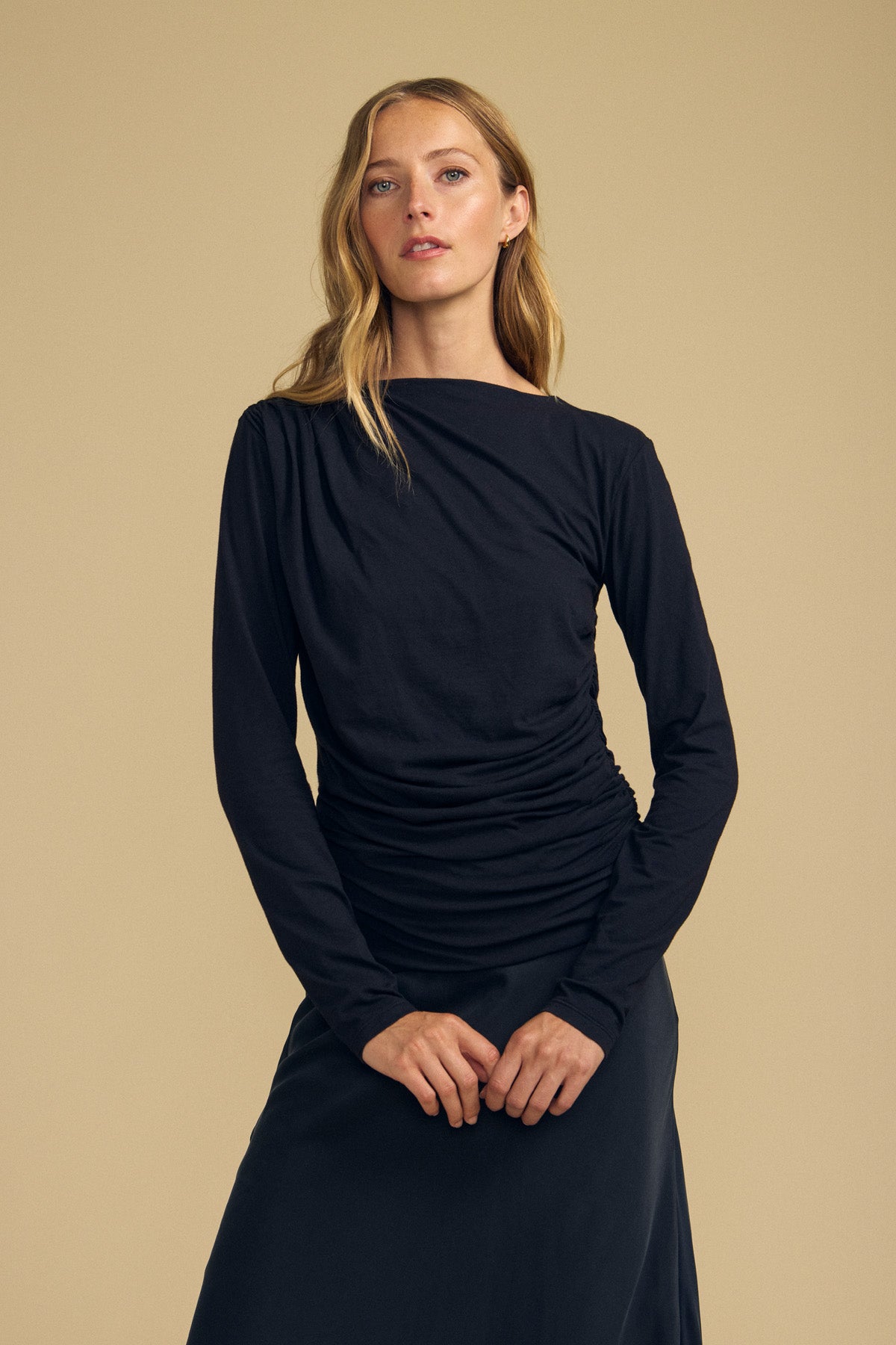 A woman with long blonde hair wears the JAZMINE TEE by Velvet by Graham & Spencer—a black fitted long sleeve top—paired with a black skirt, standing against a plain beige background and embodying everyday versatility.
