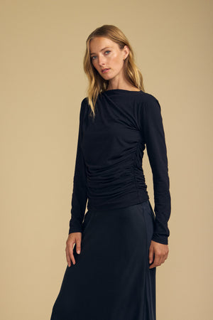 Wearing the JAZMINE TEE by Velvet by Graham & Spencer—a fitted long sleeve top made from whisper-soft gauzy fabric—paired with a coordinating black skirt, a long-haired blonde model stands against a simple beige background, showcasing an effortlessly chic look perfect for everyday versatility.