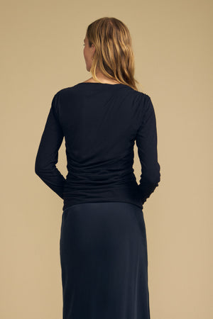A woman with long blonde hair, seen from behind, wears the JAZMINE TEE by Velvet by Graham & Spencer—a fitted long sleeve top with a boat neckline—paired with a black skirt against a plain beige background.