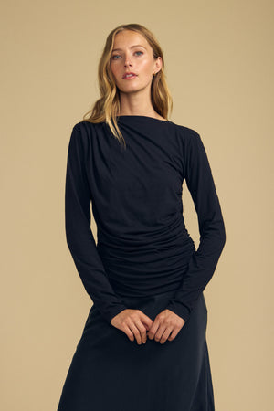 A woman with long blonde hair wears the JAZMINE TEE by Velvet by Graham & Spencer—a black fitted long sleeve top—paired with a black skirt, standing against a plain beige background and embodying everyday versatility.