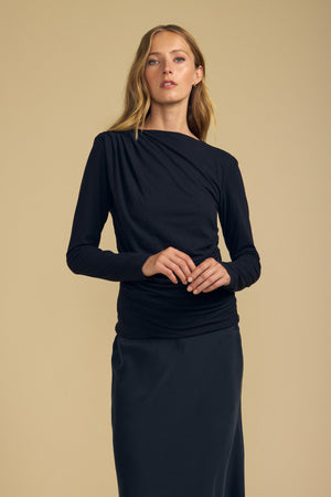 A woman stands against a beige background, wearing the JAZMINE TEE by Velvet by Graham & Spencer—a fitted long sleeve tee with a boat neckline—paired with a skirt, looking forward with her hands clasped.