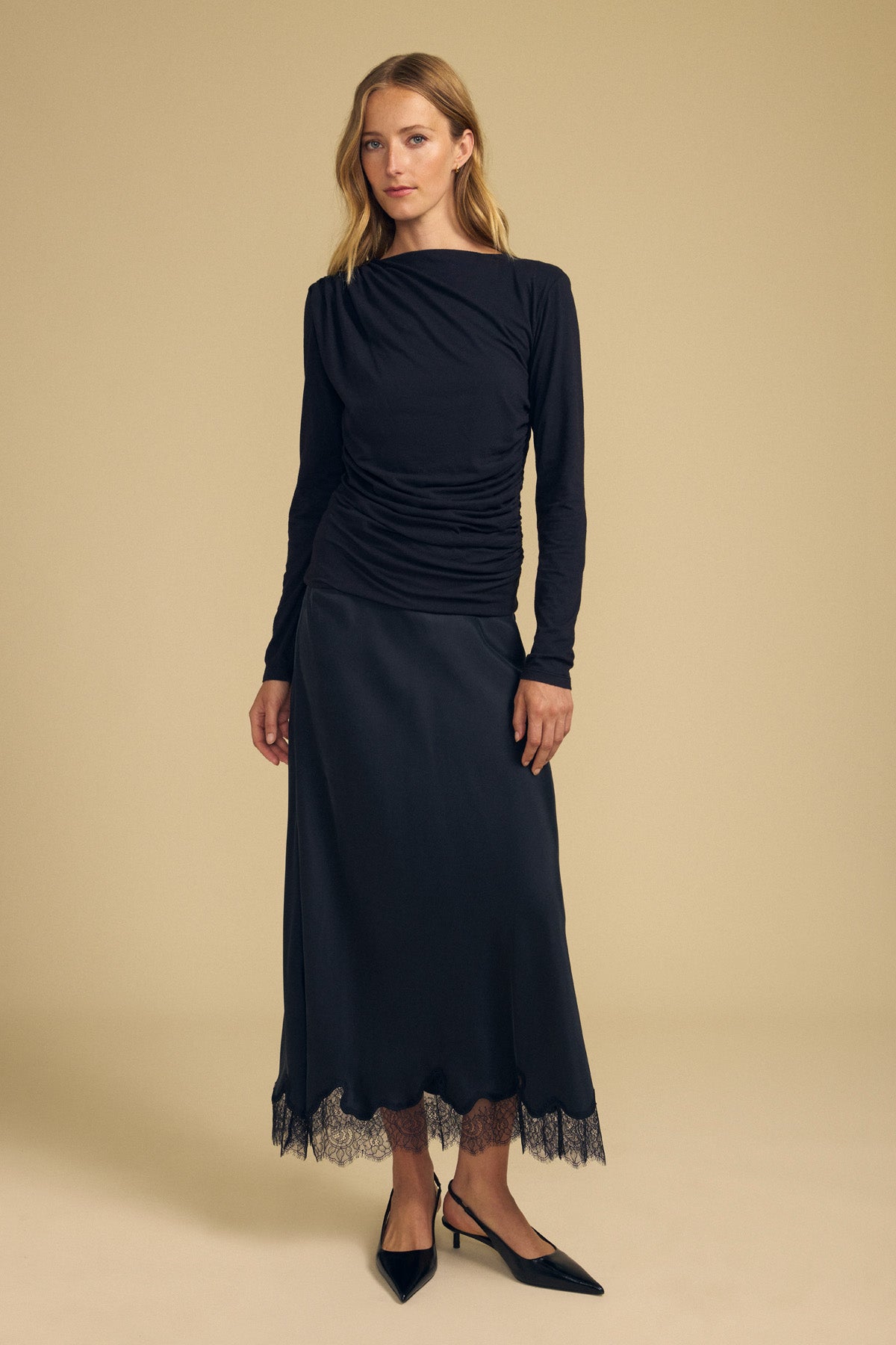 A woman wearing the JAZMINE TEE by Velvet by Graham & Spencer—a long-sleeved black top with ruched details and a boat neckline—paired with a black satin skirt featuring lace trim, stands against a beige background.-42063758819521