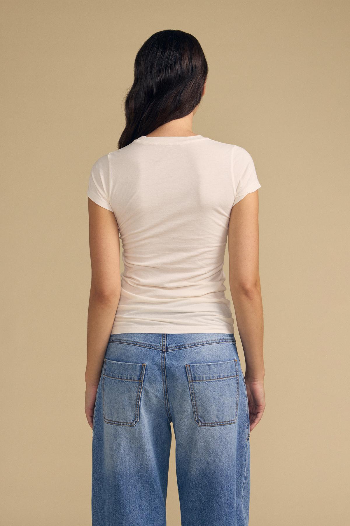A person with long dark hair, seen from behind against a beige background, wears the Velvet by Graham & Spencer JEMMA TEE—a fitted white crew neck tee—paired with blue jeans.