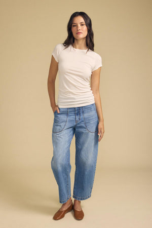 A woman stands against a beige background wearing the ultra-soft, fitted, short-sleeve JEMMA TEE in white by Velvet by Graham & Spencer, paired with loose blue jeans featuring visible stitching and brown flat shoes, with one hand in her pocket.