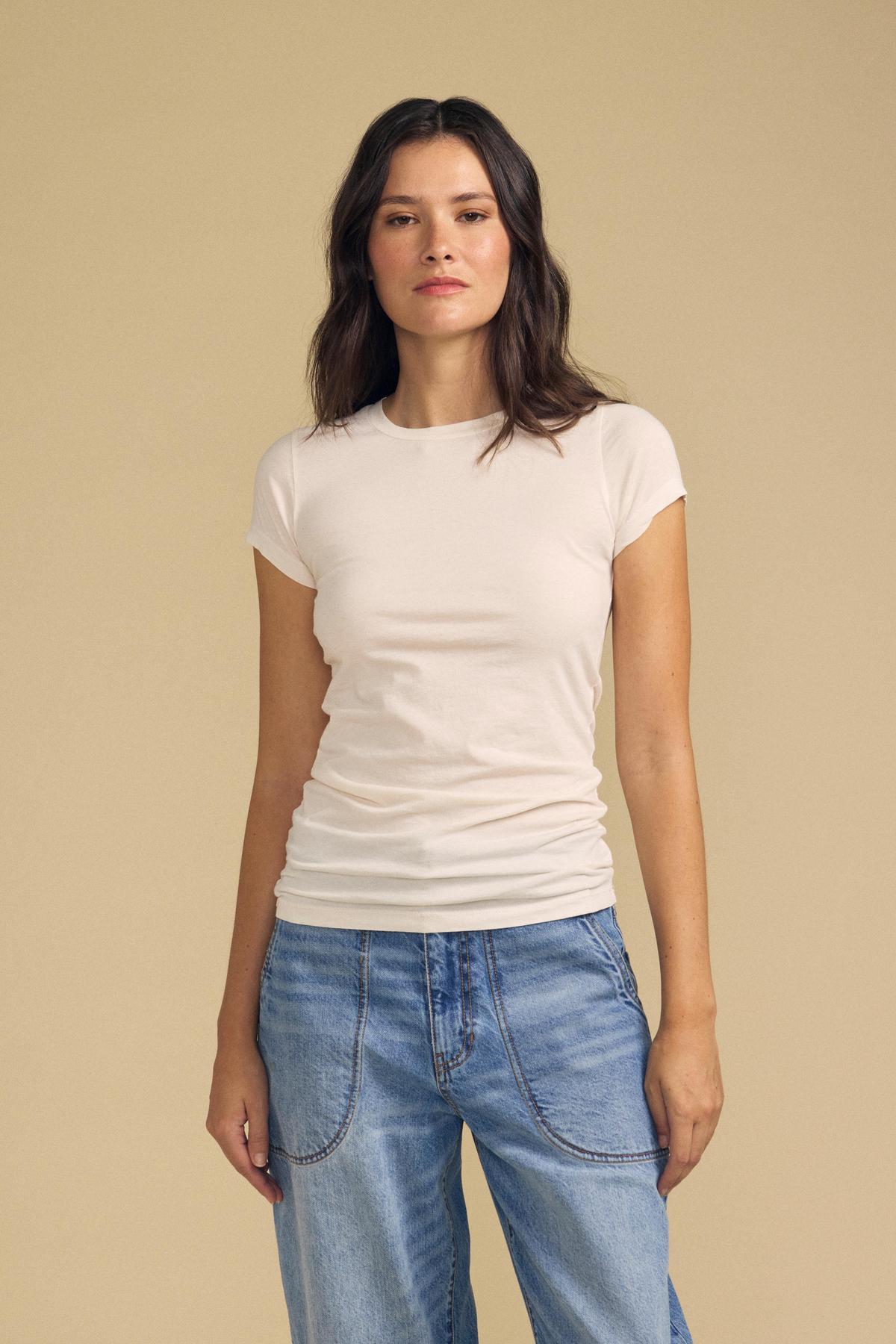 A woman with long brown hair wears the JEMMA TEE by Velvet by Graham & Spencer in ultra-soft white, paired with blue jeans. She stands against a plain beige background, facing forward with a neutral expression.-41835191828673