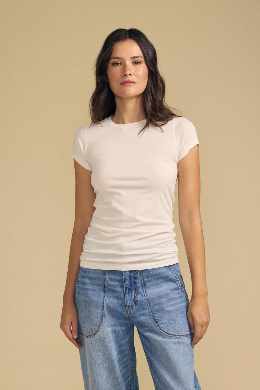 A woman with long brown hair wears the JEMMA TEE by Velvet by Graham & Spencer in ultra-soft white, paired with blue jeans. She stands against a plain beige background, facing forward with a neutral expression.