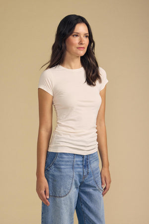 A woman with long brown hair wears the JEMMA TEE by Velvet by Graham & Spencer—a fitted crew neck tee in white—paired with blue jeans, standing against a plain beige background.