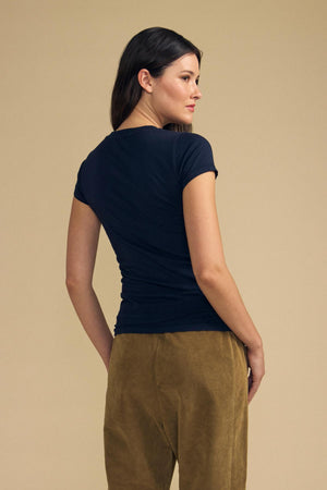 A woman stands with her back to the camera, wearing the ultra-soft JEMMA TEE by Velvet by Graham & Spencer in navy, paired with tan pants against a plain beige background.