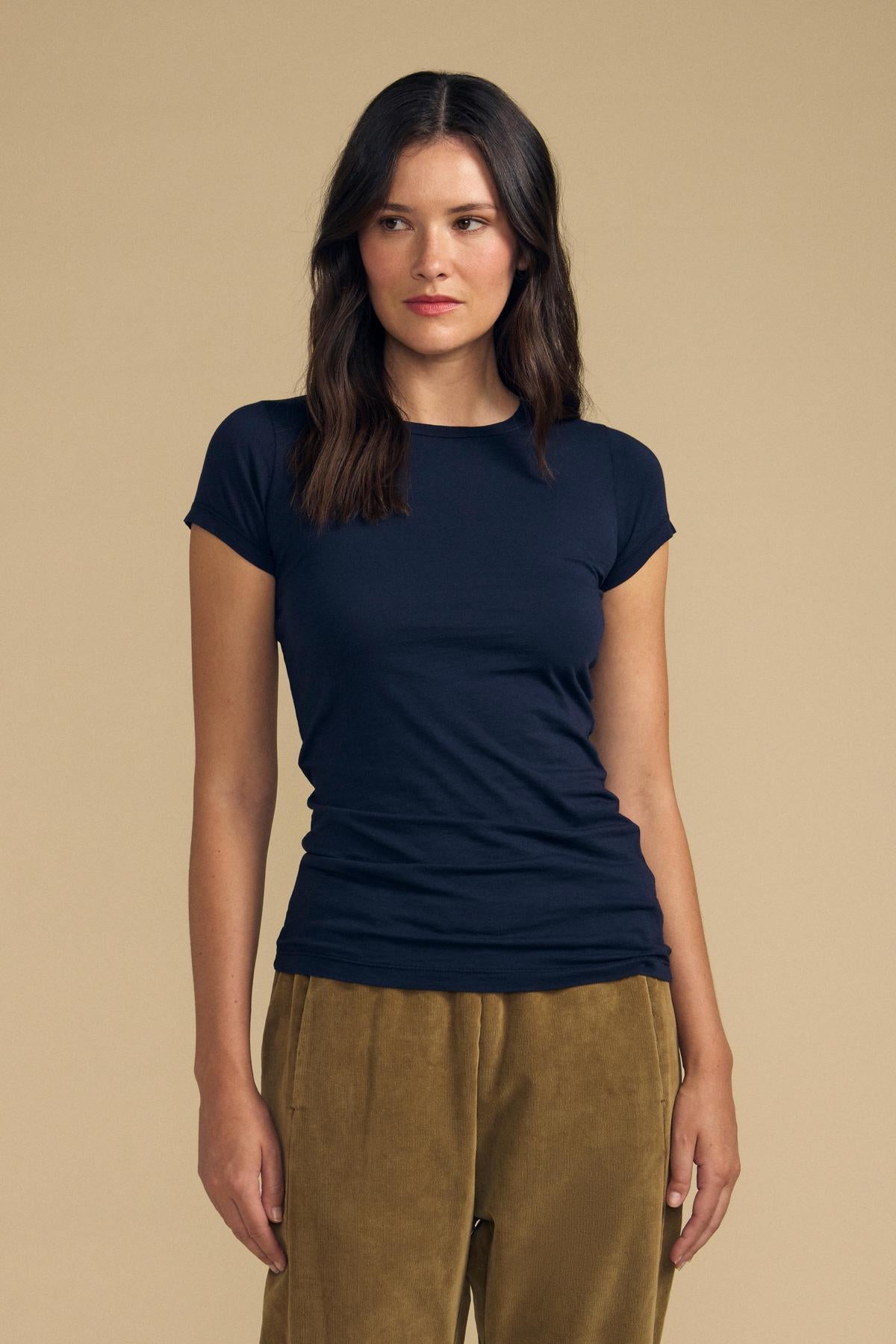 A woman with long dark hair wears the ultra-soft, fitted JEMMA TEE from Velvet by Graham & Spencer and tan corduroy pants, standing against a beige background and looking slightly to the side.