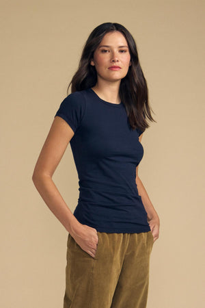 A person with long dark hair stands against a beige background with hands in their pockets, wearing the ultra-soft Velvet by Graham & Spencer JEMMA TEE in navy and tan pants.