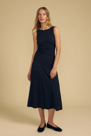 A woman stands against a beige background wearing the elegant, sleeveless black lined PIA DRESS by Velvet by Graham & Spencer and black flats, with her hair down.