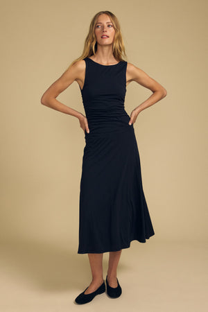 A woman stands against a beige background wearing the PIA DRESS by Velvet by Graham & Spencer—a sleeveless black gauzy whisper dress featuring an elegant round neckline and fitted waist—paired with black flats, her hands resting on her hips.