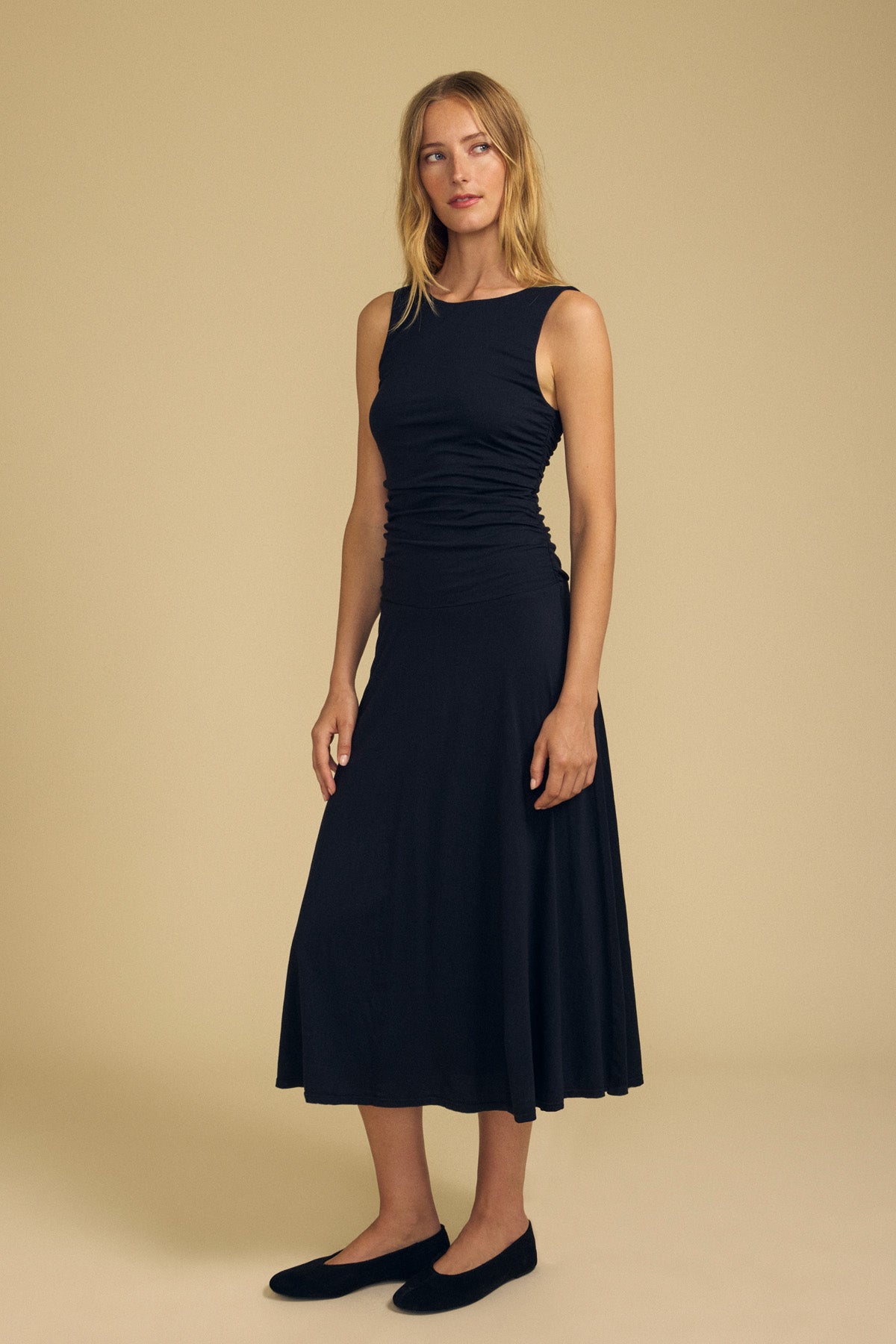 A woman stands against a beige background wearing the PIA DRESS by Velvet by Graham & Spencer, featuring an elegant sleeveless, fitted bodice and flared midi skirt, paired with black flats.-41940576960705