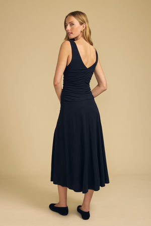 A woman stands on a beige background, facing away from the camera, wearing the PIA DRESS by Velvet by Graham & Spencer—an elegant black midi dress featuring a sleeveless design, V-back, and ruching—paired with black shoes.