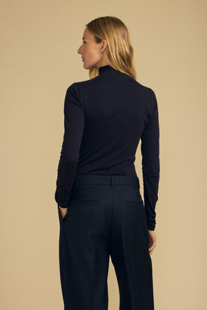 A person stands with their back to the camera, wearing the TALISIA TEE by Velvet by Graham & Spencer—a long-sleeved black mock neck—paired with black pants against a beige background.
