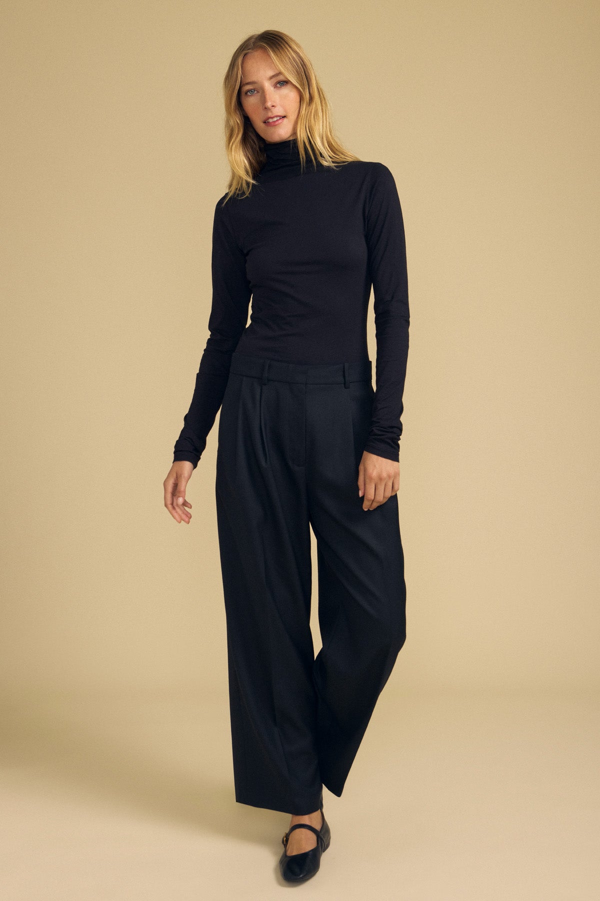 A person stands against a beige background wearing the TALISIA TEE by Velvet by Graham & Spencer—a true wardrobe staple—in black, paired with wide-leg black pants and black shoes, with their left hand slightly raised.-41964720259265