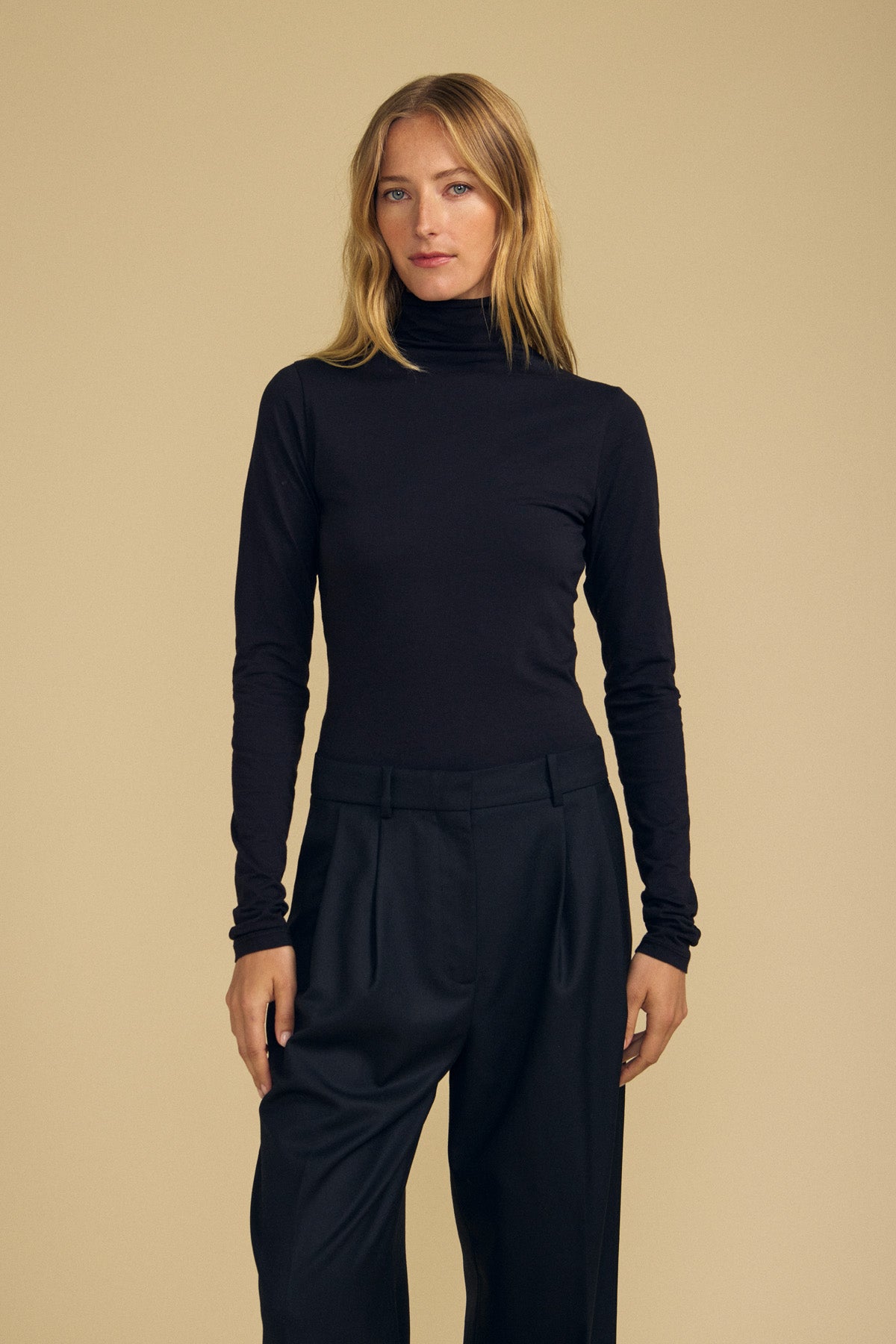 A woman with long blonde hair stands against a plain beige background, wearing the TALISIA TEE by Velvet by Graham & Spencer in black along with black pants, her expression neutral and her hands by her sides.