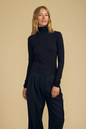 A person stands against a beige background wearing the Velvet by Graham & Spencer TALISIA TEE in black with black wide-leg pants, one hand tucked into a pocket.