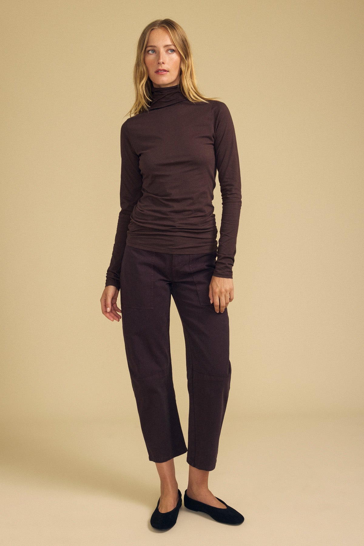 A person stands against a plain beige background, wearing the Velvet by Graham & Spencer TALISIA TEE—a long-sleeved brown mock neck turtleneck—paired with brown cropped pants and black flats.