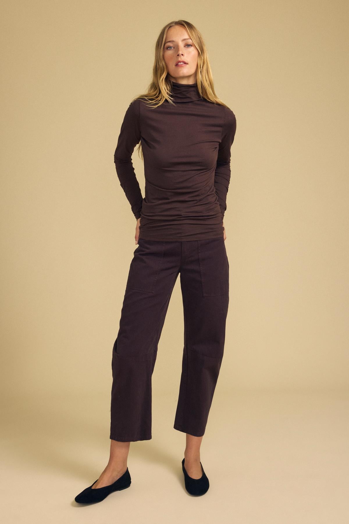 A person wearing a long-sleeved dark brown turtleneck top and BRYLIE PANT cropped barrel leg pants with utilitarian patch pockets.